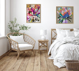Minimalist bedroom with light wooden flooring, a rattan armchair with white cushions, a small wooden side table with a white lamp, and two colorful abstract floral paintings on a white wall.