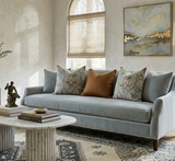 Light blue sofa with floral blue pillows and one rust-orange pillow, set in a softly lit living room with a textured rug and abstract gold-accented painting.