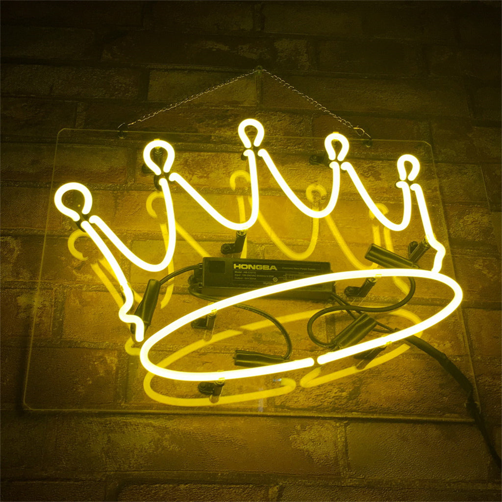 Yellow neon light shaped like a crown mounted on a clear backing, hung on a textured brown brick wall with visible electrical components.