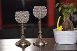Two crystal ball candle holders with metal bases on a table, next to a white pot containing a small tree and a yellow ceramic bird figurine.