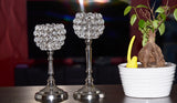 Two silver candlesticks with spherical crystal bead covers on a black surface next to a white pot with a base bonsai plant and a yellow decoration