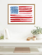Framed canvas print showing a stylized American flag in watercolor with uneven red stripes and blue field with white stars, hanging above a white shelf with plants and books.
