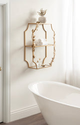 Gold-colored metal tiered shelf with a floral vine design, mounted on a white wall above a white freestanding bathtub and next to a window with sheer curtains.