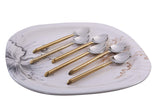 Six dessert spoons with shiny silver heads and brushed gold handles arranged on a square white plate with black and gold abstract feather-like designs.