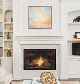 Square pastel abstract painting with soft blue, peach, and beige tones in a thin dark frame above a lit fireplace in a white built-in wall unit.
