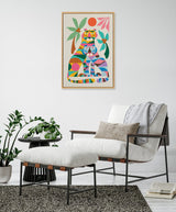 Framed canvas with colorful geometric tiger illustration featuring overlapping shapes and tropical leaves, mounted above a white cushioned armchair with ottoman and gray throw.