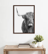 Black and white framed canvas photo of a Highland cow's head with long shaggy fur covering one eye, displayed above a wooden table with books and two potted plants.