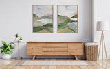 Two framed abstract landscape paintings with soft green, beige, and gray tones hanging above a wooden sideboard in a minimalist room.