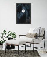 Framed canvas showing a bright full moon surrounded by swirling dark blue and black clouds, hung above a modern white cushioned armchair with matching ottoman and gray throw in a minimal room.