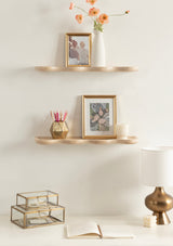 Two wall-mounted wooden shelves with curved edges holding framed photos, a tall white vase with orange flowers, a small gold geometric pencil holder, and a white candle, above a table with glass boxes, an open book, and a brass-ac