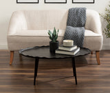 Oval black coffee table with scalloped edge and three legs in front of a light beige curved-back sofa with a gray knit throw draped over one arm.