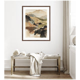 Framed abstract watercolor painting of layered mountains in beige, brown, black, and green tones, hung above a rustic wooden bench with a book and a draped light fabric.