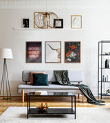 A modern living room with a light gray sofa adorned with dark green and pink pillows, a dark green throw draped over the arm, and three framed artworks above: a pink neon 'Good Vibes Only' sign, a highland cow portrait, and a dark