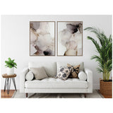 Two framed abstract canvas art pieces with soft brown, beige, black, and gray smoke-like patterns above a white sofa with patterned pillows in a bright living room.