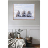 Framed canvas print of three snow-covered pine trees against a pale sky, hanging above a beige textured blanket on a couch and a small wooden side table with a white mug.