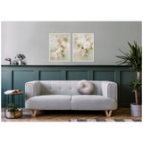 Two framed abstract floral paintings in soft pinks and greens above a modern light gray sofa with round cushion, flanked by a side table and potted plant against green-gray paneled wall.