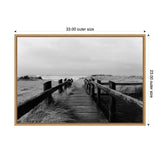 Black and white photograph of a wooden beach boardwalk bordered by grass, leading toward the ocean horizon under a cloudy sky, framed in light wood.