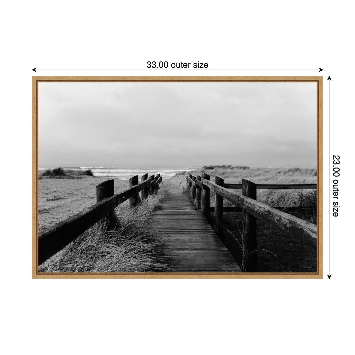 Black and white photograph of a wooden beach boardwalk bordered by grass, leading toward the ocean horizon under a cloudy sky, framed in light wood.