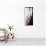 Vertical abstract canvas wall art with swirling black, gold, white, and gray tones in a black frame, hung on a white wall above a wooden side table with a vase and fern.