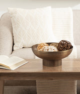 Dark bronze ribbed metal bowl on wooden coffee table holding four decorative woven and natural balls, with light beige sofa and textured pillows in the background.