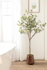 Tall faux eucalyptus tree with green round leaves in a copper-colored cylindrical pot, placed on a light wood floor beside a white bathtub and sheer curtain.