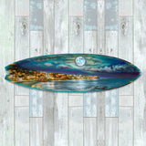 Long surfboard-shaped wall art depicting a nighttime coastal scene with a full moon reflecting over a calm ocean and illuminated shoreline houses.