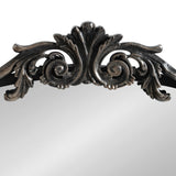 Close-up of dark metal ornamental carving with silver highlights forming an arch at the top edge of a mirror frame, featuring scroll and leaf motifs.
