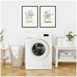 Minimalist laundry room with white front-loading washer, two framed botanical line drawings in black frames, tan wicker laundry basket, and white table with green plant and detergent.