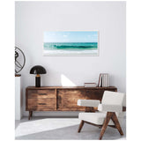 Framed canvas of a calm ocean wave under a blue sky with light clouds, hung above a mid-century modern wooden sideboard and beige armchair in a minimalist room.
