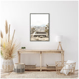 Framed canvas print of a beige vintage van parked in a Joshua Tree desert landscape with mountains in the background, hanging above a rustic wooden console table with pampas grass and wicker decor.