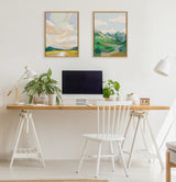 Minimalist home office with a wooden desk on white trestle legs, two abstract landscape paintings with pastel and green tones hung above a black computer monitor, and potted plants on the desk.