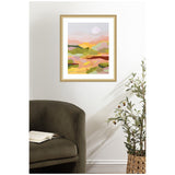 Abstract landscape art with layered pastel hills in pink, green, and orange tones, framed in light wood and hung on a white wall above a dark green upholstered chair with a book open on it.