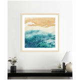 Abstract square artwork with warm sand tones blending into deep blue and teal waves, hung above a black console with a lamp, plant, and books.