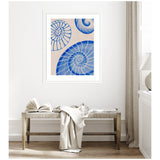 Framed artwork featuring three blue spiral shell designs on a beige background, hanging above a light wood bench with an open book and folded textiles.
