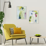 Two abstract paintings with green, blue, and purple hues in white frames hung on a beige wall above a yellow armchair and round wooden table with green apples.