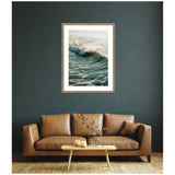 Framed wall art print of a breaking ocean wave with soft morning light, mounted on a dark gray wall above a brown leather sofa and a wooden coffee table with a clear vase.