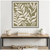 Framed square canvas with cream silhouette botanical leaves on olive green background, hung above a wooden table with books, a white vase with greenery, and a beige mug.
