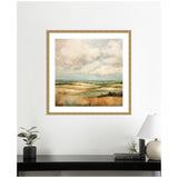 Framed abstract landscape painting showing rolling hills in muted green, tan, and blue tones beneath a cloudy sky, hung on a white wall above a black console with plants and a lamp.