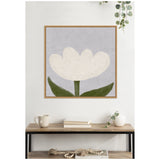 Minimalist white tulip with three green leaves on a muted gray background, framed in light wood and hung above a wooden table with books, a cup, and a white vase.