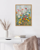 Framed vertical floral painting with colorful wildflowers in yellow, red, pink, and white on a muted green and blue background, hanging above a white desk and vase with pampas grass.