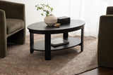 Oval dark wood coffee table with lower shelf holding books, topped with a beige ceramic vase with green leaves and a small black box, set on a textured brown rug between two green upholstered chairs.