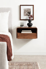 Dark wood floating side table with an open shelf and drawer, holding a black vase with dried flowers and a gray candle, mounted beside a bed with a white pillow and brown knitted throw.