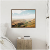Framed canvas featuring a panoramic landscape of rolling green and brown hills under a partly cloudy blue sky, hanging above a rustic wooden bedside table.