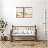 Two framed pastel landscape paintings with soft greens, pinks, and blues hang above a wooden bench with white cushions, flanked by a woven basket plant and a lantern on a hardwood floor.