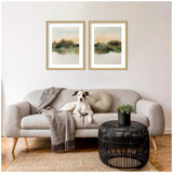 Two framed abstract landscape prints with soft green, beige, and brown tones hanging above a light grey curved sofa with a grey throw, round cushion, and a small black woven coffee table with a plant.