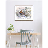 Watercolor painting of a covered wooden bridge surrounded by snow and trees, framed and hung above a minimalist desk with a plant and metal decor.