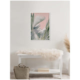 Framed canvas of green Bismarck palm leaves against a soft pink background hanging above a white bench draped with a cream fur throw, next to a beige bag and dried pampas grass.