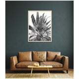 Black and white framed print of palm fronds hangs above a brown leather sofa with a wooden coffee table and vase against a dark gray textured wall.