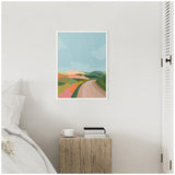 Abstract landscape artwork with a winding road leading through overlapping hills in pastel pinks, greens, and blues, framed and hung on a plain light wall above a rustic wooden bedside table.