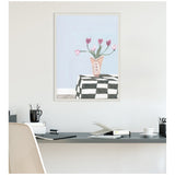 Framed canvas wall art showing a pale pink vase with tulips on a black and white checkered table against a light blue background, hanging above a minimalist desk setup.